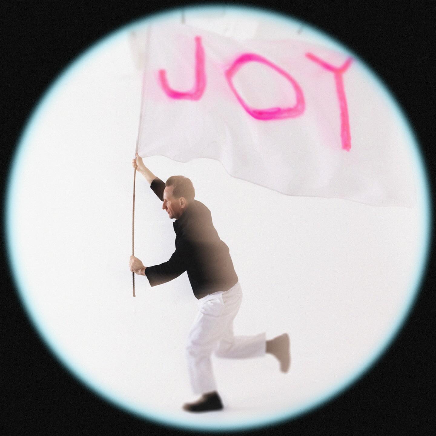 Album Art of Martin Smith's Joy album - On a black background, a white circular hole with a slight blue tint on the edges is showing a picture of a middle-aged man in white pants and a black coat running and holding a large, white flag that has the word 'Joy' hand-drawn on it in pink marker.