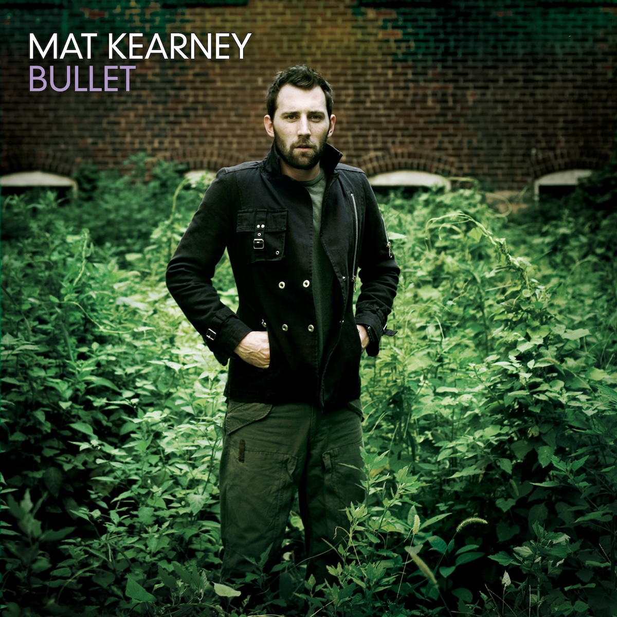 Album Art of Mat Kearney's Bullet album - A photo of a young man with a beard and short brown hair wearing green cargo pants and a green T-shirt with a black, double-breasted coat that is open slightly has his hands in the pockets of the coat. Behind him and around his feet is green foliage of some leafy plants, a bush or something. The plants are covering up the windows in a brick wall in the background. In the top left, printed in large white text is the artist name, and below it in purple the album title.
