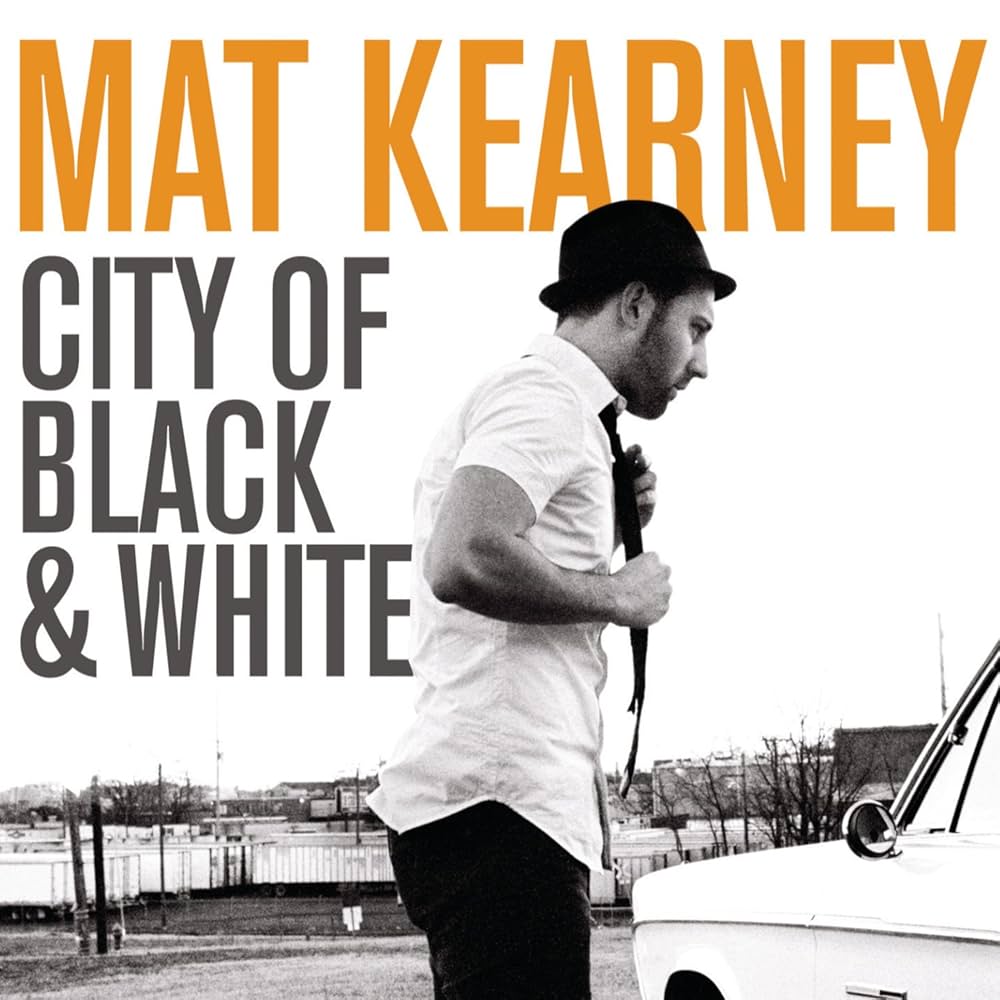 Album Art of Mat Kearney's City Of Black And White album - A black and white photo where the sky is white, and the man stands next to a white car, adjusting his black tie. He's wearing a black hat, a white shirt, and black pants. Behind him is some one-story buildings, tractor trailers, and poles along with trees. Above his head, the artist's name is printed in large orange letters. To the left of him, the album title in smaller black letters.