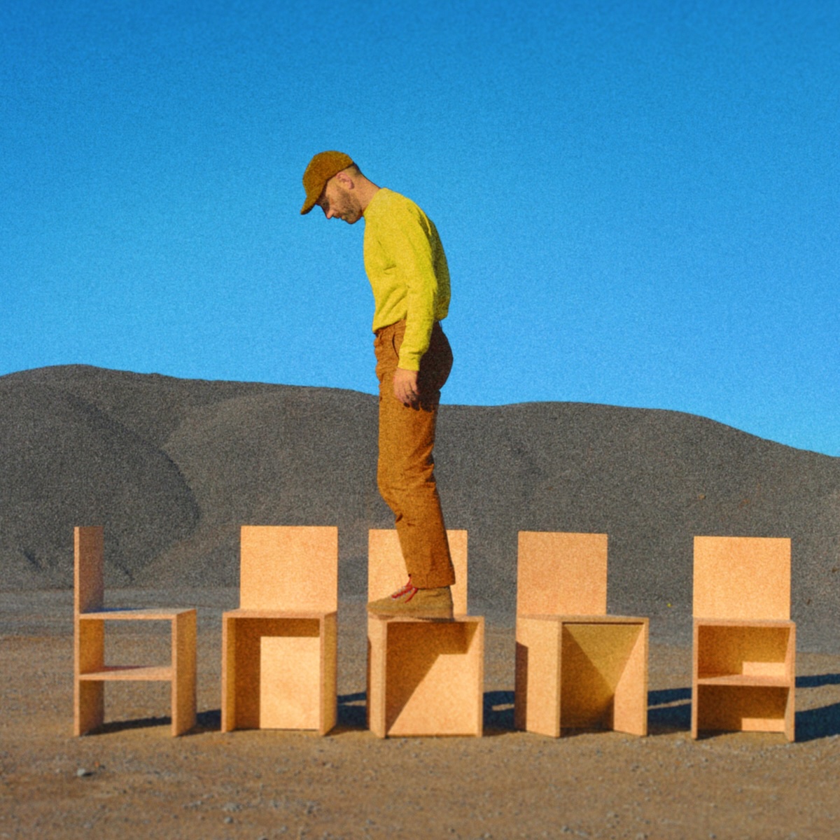 Album Art of Mat Kearney's January Flower album - On what looks like a flat, dirt surface with a blue sky and a rolling hill in the background, a grainy photo of a man and 5 chairs. The wooden chairs of a light brown color, and they are a very simple box to sit on with a very rectangular wooden back. Each of the chairs has a bit different design below the seat. A white man with short hair and a baseball cap is in a yellow shirt and orange pants, standing on the middle chair and looking down in a profile pose.