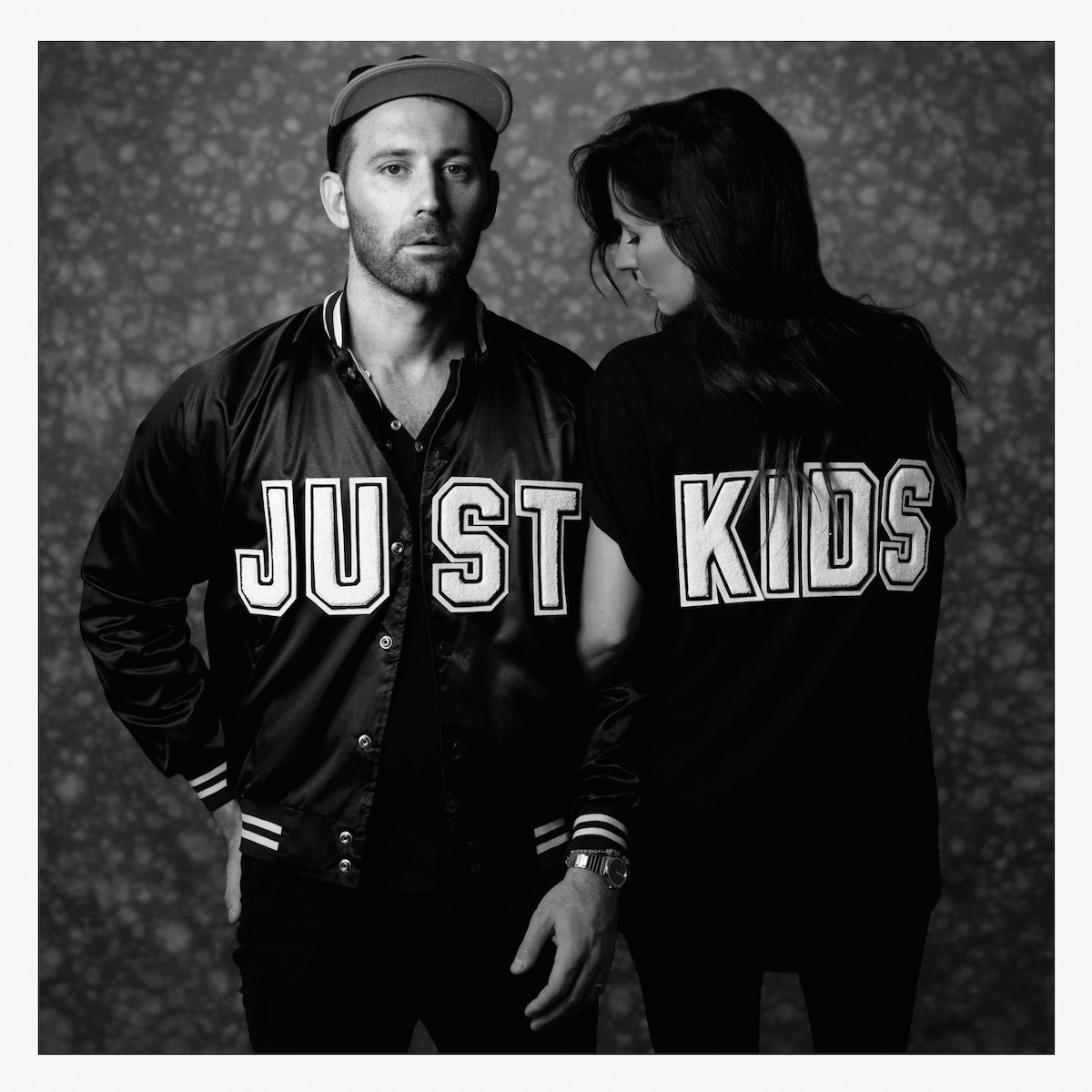 Album Art of Mat Kearney's Just Kids album - A black and white photo of a man standing next to a woman. The man is on the left, wearing a black letter jacket with the letters 'JUST' on the front and his hands at his sides. He's looking at the camera with a bit of a blank expression. The woman has long, dark hair and has her back to the camera and is looking off behind the man. Her arm is interlocked with the arm of the man. She's wearing a black shirt with the letters 'KIDS' in the same letter style as the letter jacket. The background is gray and mottled, and there's a thick, light gray border around the edge.