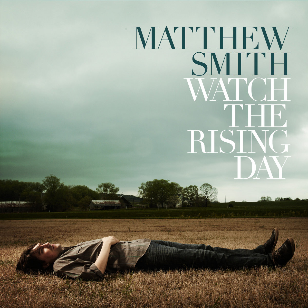 Album Art of Matthew Smith's Watch The Rising Day album - In a field of brown grasses, a photo of a man laying down in the grass and looking up into the green, cloudy sky is shown. In the background looks to be some trees and some farm buildings. and the top half is all cloudy sky. The person, a white man with fairly short hair, is wearing dark pants and a brown shirt and has right hand over his stomach. Printed over the sky is his name in a dark green color, and in white below it is the album title.