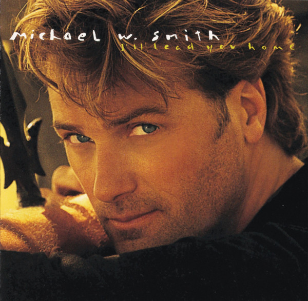 Album Art of Michael W. Smith's I'll Lead You Home album - A photograph of a young, white main with stubble and tousled brown hair looking over his shoulder into the camera, and leaning up against a fence or something. It's a very close-up photo of his face, so not much can be seen of the background or the black shirt he's wearing. Towards the top, in white in a hardwriting-style font is the artist's name, and below it in smaller yellow font is the album title, slightly off to the right.