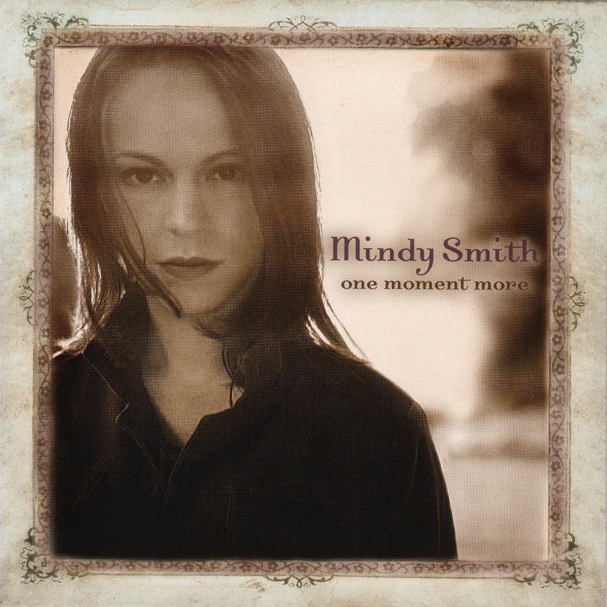 Album Art of Mindy Smith's One Moment More album - It looks like it's a photo of a young woman with shoulder-length hair mostly lit from behind. The background is out of focus, but it looks like just a field with open sky and maybe some trees off to the right. Around the photo is what looks like an illustration that looks like a photo frame, with a decorative brown border around the photo and then green leaf details in the middle and corners of the edges on a wider beige border.