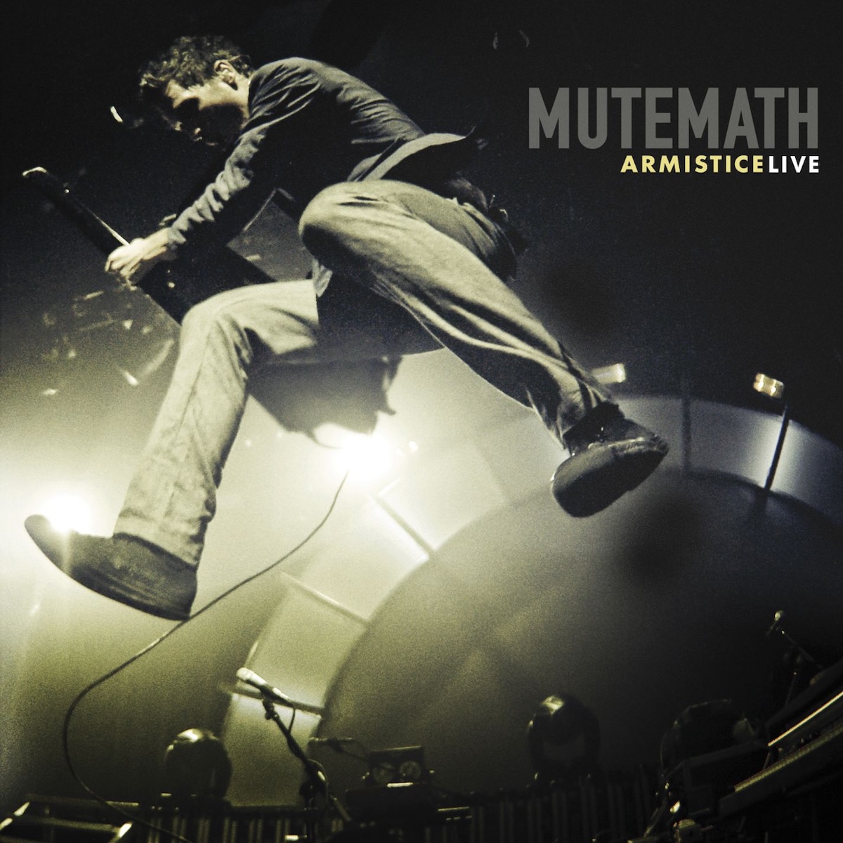 Album Art of Mutemath's Armistice Live album - A black-and-white photo where the white parts has a bit of a gold hue as well. The background is mostly dark and we can see that the picture is taken from the ground looking up. A man is jumping in the air and the closest thing to the camera are his feet. He's towering above the camera and also playing a keytar. Behind that, we can see the bright lights of a stage and an arched backdrop, with a few microphones and lights on the stage in front of it. In the top right, the band name is printed in dark grey. And below it in a light yellow is the word 'Armistice' and then 'Live' in white.