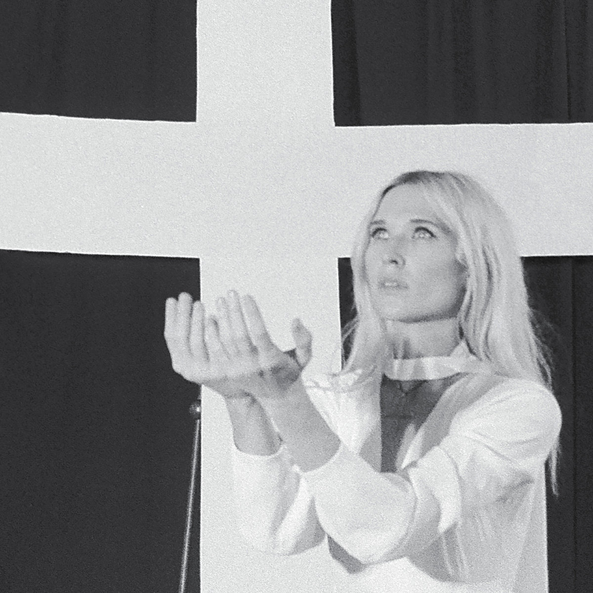 Album Art of Natalie Bergman's Mercy album - A grainy black-and-white photo of a white woman with blonde hair standing in front of a black background with a large white cross. The woman is wearing a white coat with a V-neck and a strap across the neck, and her arms are held out in front of her together, palms facing up. She is looking up towards the sky in the direction of the light, as the hands strike a shadow on her chest.