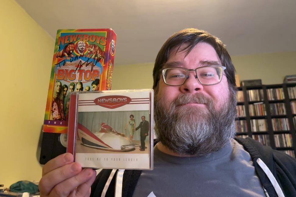 A photo of a white man with brown hair and a graying beard and glasses looking slightly down at the camera, smiling, and holding to the left of him a few items. First is a CD of the Newsboys album in a case, then the second is a VHS video peeking out from a paper slipcase. The VHS case says 'Down Under The Big Top' and 'Newsboys' and is autographed by Steve Taylor.