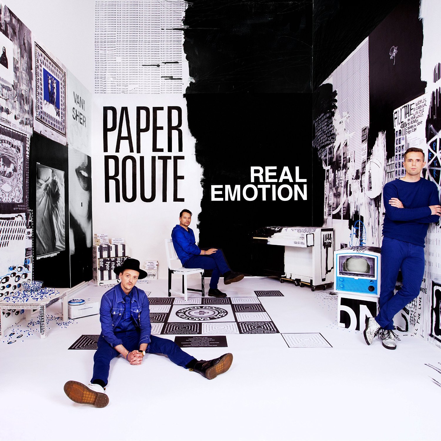 Album Art of Paper Route's Real Emotion album - A photo of what looks like a square room, although a closer look shows that the back wall keeps going up and there's no ceiling visible in the room. The left half of the back wall is white, and the right half is black, and the area where they meet is not a straight line, but more a haphazard edge. Next to the black wall is a piano, and half of it is also black and the other half white with an illustration painted on the side of it. The white walls have graphics or even handwriting in black or color on them, though the majority of the color is black, white or blue. Three men are scattered around the room, one standing against the right wall, one sitting on a chair in the back, and one sitting in the middle of the floor. They're wearing dark blue clothes.