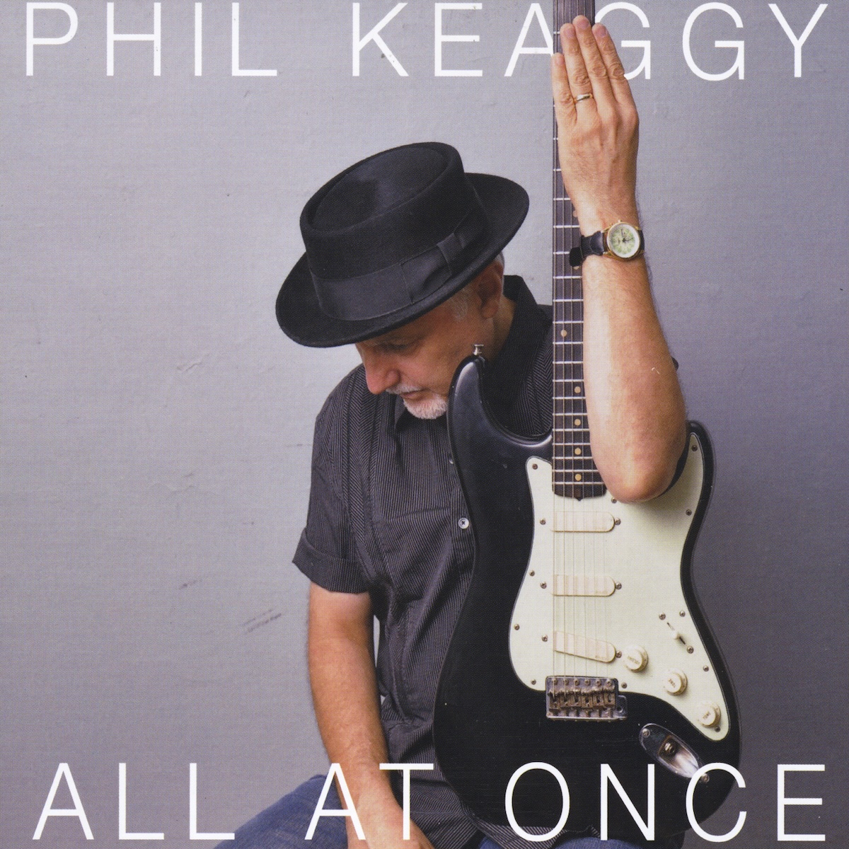 Album Art of Phil Keaggy's All At Once album - In front of a gray wall, an older man with grey hair and a black top hat on is wearing a black shirt and holding a black electric guitar with white around the pickups. He has the guitar sitting on his knee and standing vertically above his head, with his arm extending above his head up the fretboard. He's looking down in front of him and has a slight smile on his face. The artist's name is printed across the top in large, thin white letters. And the album title in slightly smaller letters across the bottom.