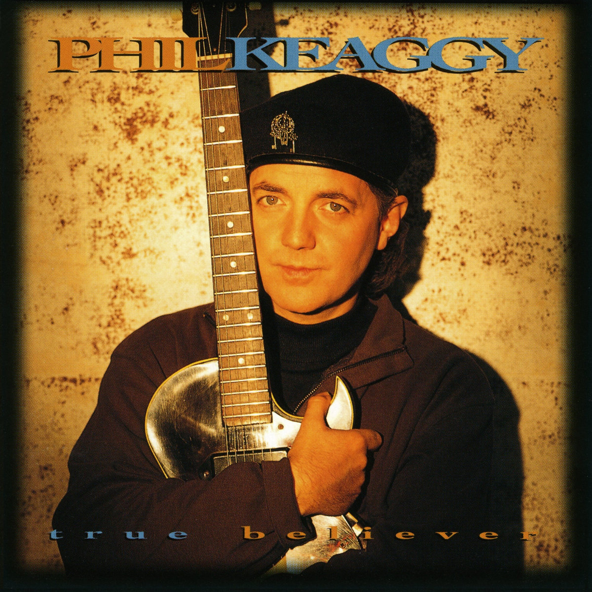 Album Art of Phil Keaggy's True Believer album - A photo of a middle-aged white man holding a shiny, silver guitar in an embrace, his arms around the body of the guitar and the neck up next to and above his head. The man is looking at the camera with a pretty blank expression and is wearing a brown jacket over a black turtleneck and a black beret on his head. He is bathed in a warm, orange-ish light, and behind him can be seen a beige wall which might be rusting in places or something, as there's dark brown marks in many places. At the top, the artist's name is printed in large letters, first name in orange and last name in blue. At the bottom, in much smaller letters, is the album name, first word in blue and the second word in orange.