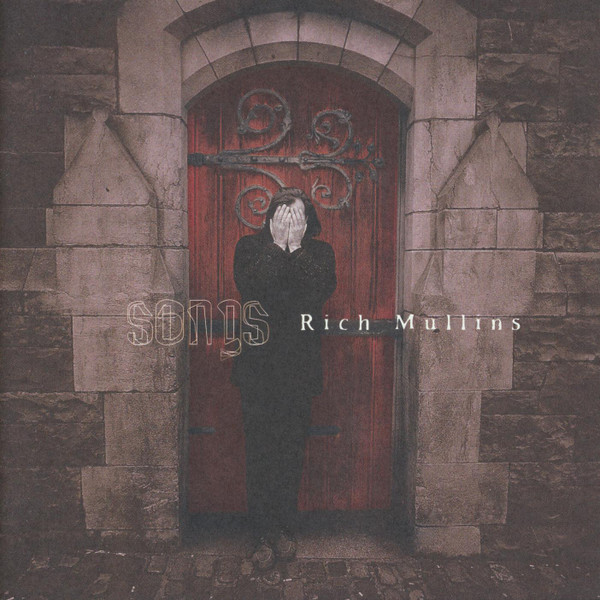 Album Art of Rich Mullins's Songs album - A photo of a man standing in front of an old stone doorway with a wooden door that has a red hue and some ornate black metalwork coming from its hinges. The man has shoulder-length brown hair and is wearing black. He's facing towards the camera, but has his hands completely covering his face. The outline of the word 'songs' is printed over the image, and in a smaller white font the artist name is printed to the right of that.