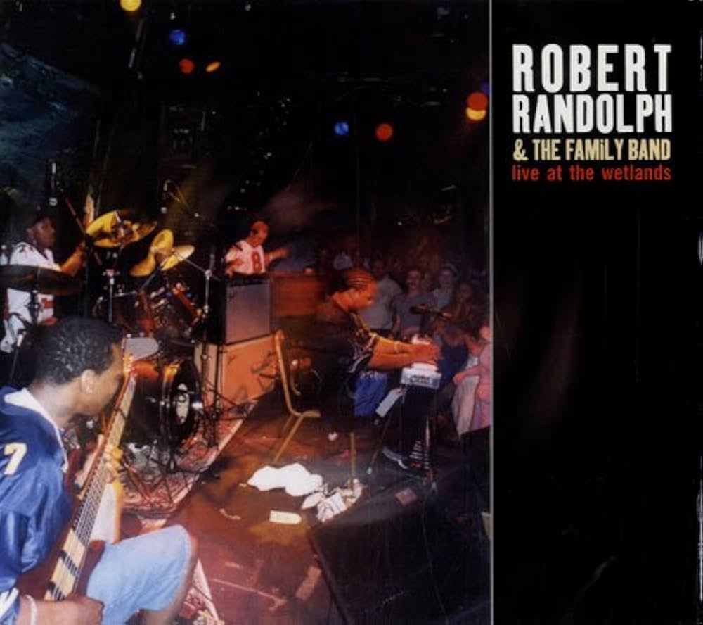 Album Art of Robert Randolph & The Family Band's Live At The Wetlands album - On the right side, the band's name and the album title is printed on a black background. On the left two thirds is a photo of the band on stage, a black man in a sports jersey sitting in the left foreground playing a bass guitar, and drums and keyboards behind him. Between the drums and the keys are a few amps, and in front of those is Robert Randolph sitting and playing a pedal steel guitar. To the right of him, the crowd can be barely seen watching the show in the dark room.