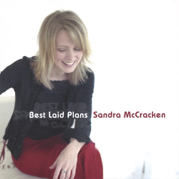 Album Art of Sandra McCracken's Best Laid Plans album - On a white background, a young white woman with shoulder-length blonde hair is sitting in white chair wearing a black sweater and shirt and red pants. In front the photo of her is printed the album title in white, and besides that, her name in a dark red that matches the pants.