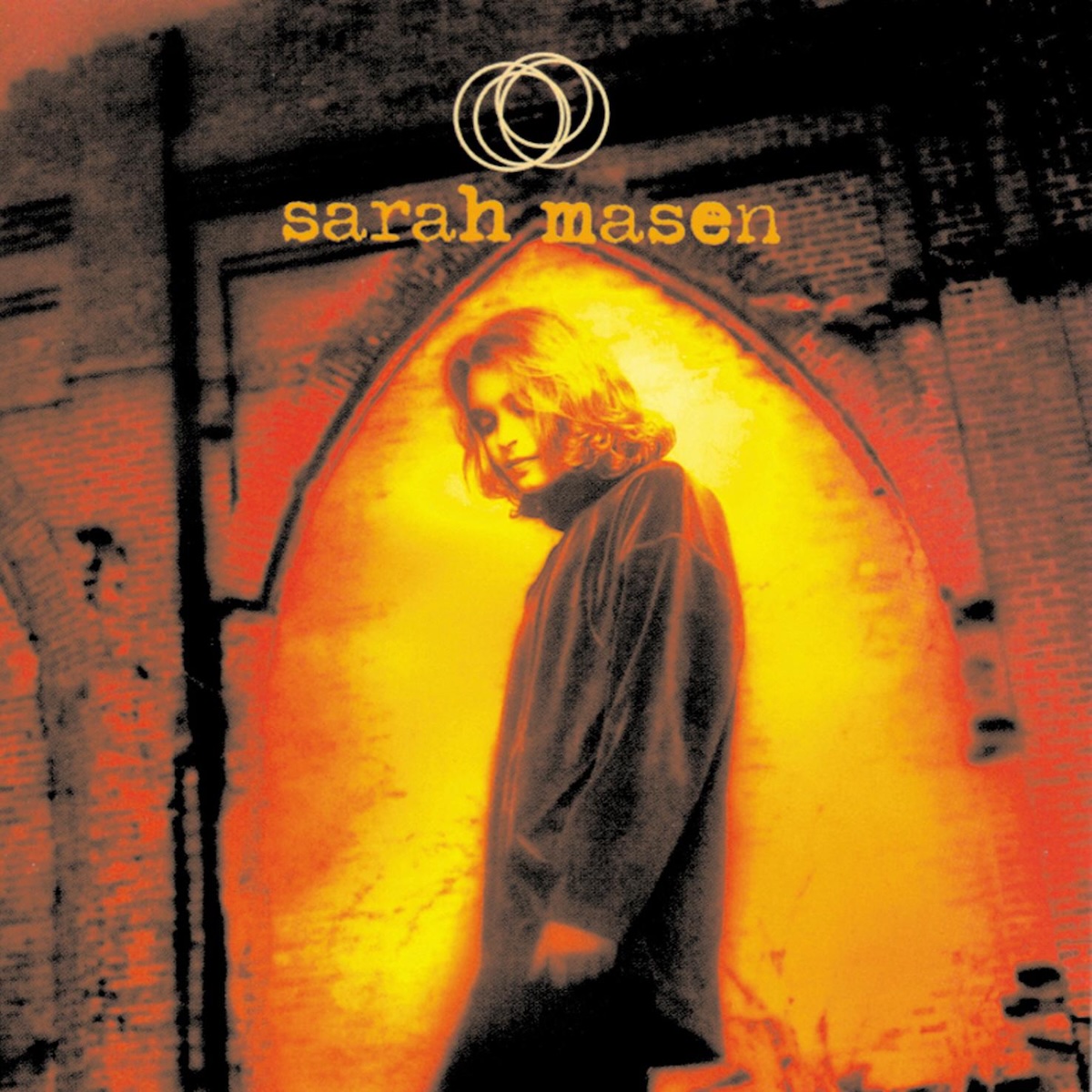 Album Art of Sarah Masen's self-titled album - An over-exposed or oddly edited photo of a young woman taken from closer to the ground in front of her. She's looking down with her eyes mostly closed and has a serene look on her face. Her hair is down to her shoulders and she seems to be wearing a dark coat. Her face and wall behind her is a gold color, while some of her clothes and the brick wall behind her are red, then brown and black, so maybe it's some sort of gradient of colors replacing the actual colors. The background seems to be a large brick wall a bit behind her, with a brick arch built into the wall, and she's framed inside the archway. Above her head is printed her name, and there's four interlocking circles printed above that.