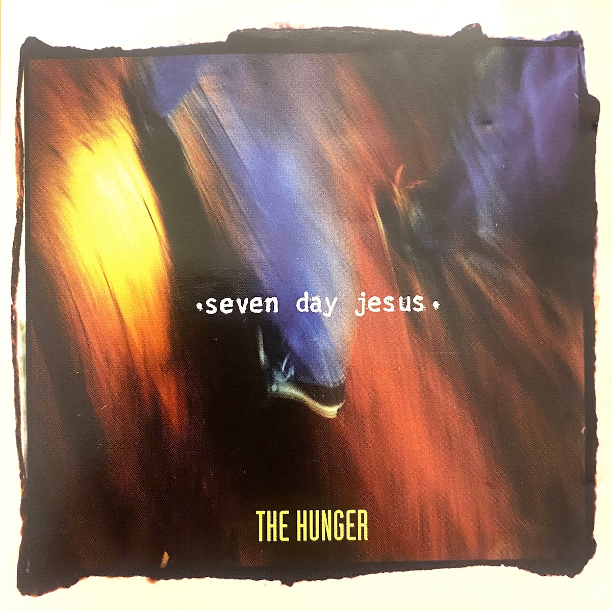 Album Art of Seven Day Jesus's The Hunger album - On an off-white background, a grainy photo of some colors of yellow, blue and red are shown. It can't tell if it's a blurry photo of some paint, or maybe even outer space or something, it's just so distorted and blurry. Around the photo is a very rough, black background which seems hand-drawn. In the very middle, on top of the photo is printed the band's name in white, all-lowercase text in a typewriter font. And towards the bottom of the photo, the album title is printed in a thinner, taller font in yellow.