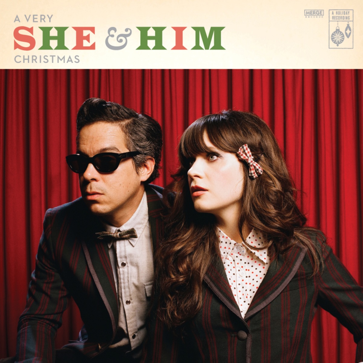 Album Art of She And Him's A Very She And Him Christmas album - The bottom four fifths is a photo of a white man and white woman from the chest up wearing black suits and white-colored shirts beneath. They are in front of a red curtain and seem to be looking at something to the left of frame with some awe or surprise. The man has a bowtie and is wearing sunglasses. At the top in a golden white background it has written 'A Very She And Him Christmas', with the band name much larger and in green and red with a grey ampersand. In the top right, smaller items say 'A Holiday Recording' and the Merge Records logo is also there too.
