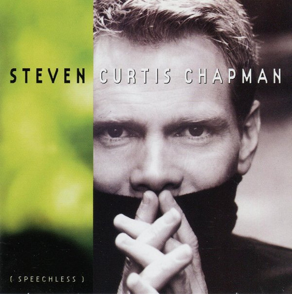 Album Art of Steven Curtis Chapman's Speechless album - On the left side, the background is a blurry green and gold, maybe a blurry shot of some bush or something, fading to black at the bottom. On the right two thirds is a somewhat desaturated photo of a middle-aged man with short, light hair wearing a turtleneck with the collar covering up his mouth. The photo is just from his shoulders up, and he has his fingers in a praying pose in front of his covered mouth and is looking directly at the camera. Across the top, it has the artist's name, with his first name in black over the green background, and his 2nd and last names in white above the photo. In the bottom left corner, in white on the black is written '[ Speechless ]'.