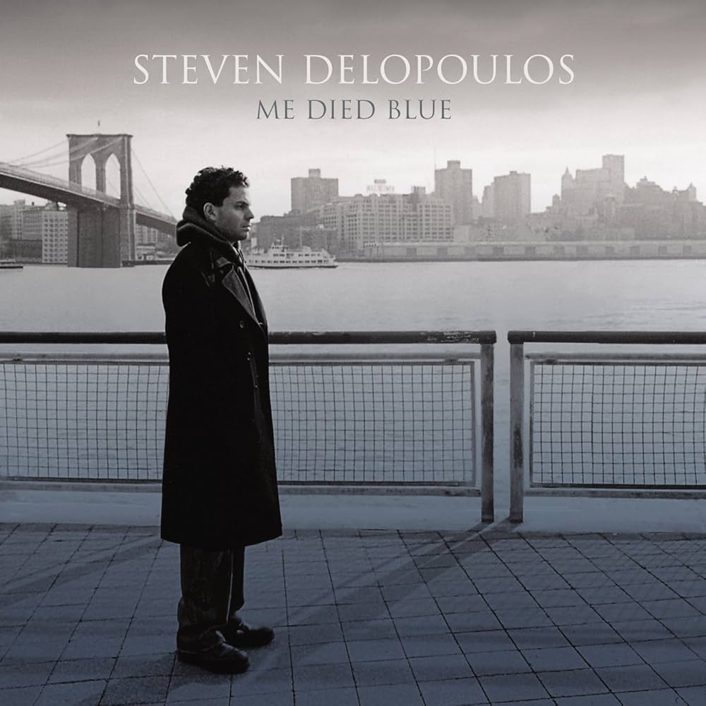 Album Artwork of Steven Delopoulos's Me Died Blue - Standing on a walkway with a railing behind them, a man in a long black jacket is facing to the right. Behind him, a wide river stretches and a ferry boat can be seen on the other bank, along with lots of buildings of the New York City borough of Brooklyn. Off the left side can be seen the Brooklyn Bridge. In the gray sky is the artist name in white and the album name in a darker gray.