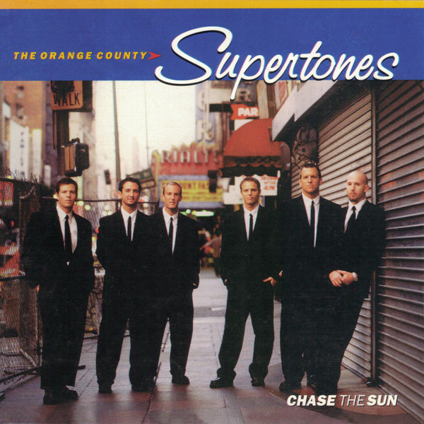 Album Art of The O.C. Supertones's Chase The Sun album - Six young men are photographed standing on a city sidewalk with a theater marquee behind them and a metal door on the front of a shop off to their right. One of them is leaning against the metal door. They are all wearing white shirts and black ties, and suits and black pants that look a bit baggy. They're all looking at the camera with mostly blank expressions, a few slightly smiling. Above the photo the top fifth vertically is a bright blue, with 'The Orange County' printed in small, orange text, and then 'Supertones' printed in much larger white text in a stylized cursive-ish font. At the bottom of the photo it has 'Chase The Sun' printed in smaller, uppercase letters in the bottom right.