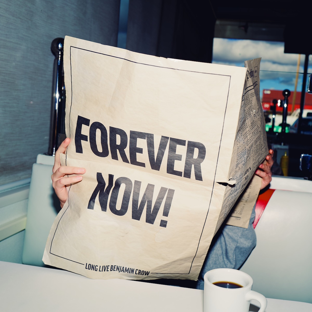 Album Art of Switchfoot's Forever Now album - A photo of a man reading a newspaper at a diner booth that is white. He's got in front of him a newspaper, where we can only read the back page design, which says 'Forever Now' in huge capital letters, and has a border which is interrupted in the bottom middle by the words 'Long Live Benjamin Crow'. The paper is clearly thin and has been torn in a few spots where it has been folded. All we can really see of the man is their hands holding the sides of the paper, and a power blue sleeve covering their left arm. The person has coffee in a white mug in front of them on the table.