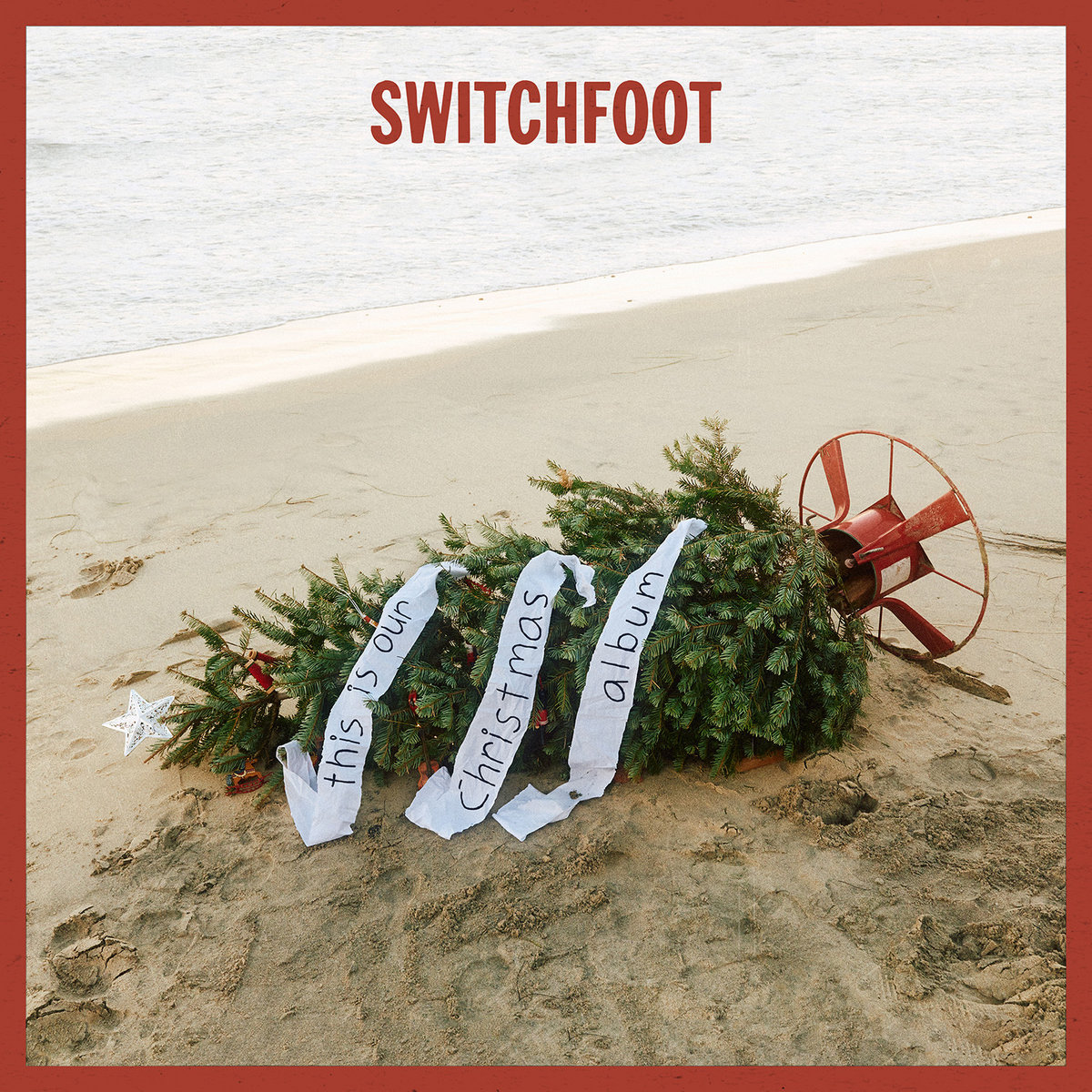 Album Art of Switchfoot's This Is Our Christmas Album album - A photo of a fir tree decorated with a star and tinsel and a banner that has written on it 'this is our Christmas album' that is on a beach and laying on its side in the sand, with the red tree stand attached to the bottom on the right. Behind the sandy beach can be seen ocean waves. At the top, in a dark red, is printed the band name. A dark red border is also around the photo.