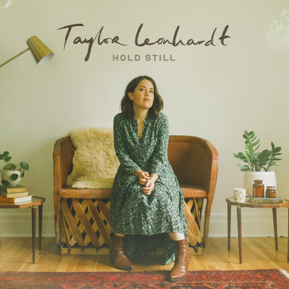 Album Art of Taylor Leonhardt's Hold Still album - A woman sitting in a room on a wooden loveseat with leather seating, it looks to me. The wall behind is light green, and on both sides of the loveseat is endtables with 3 books under a small potted plant on the left, and one book in front of a larger potted plant, with a mug and some decorative glass jars on top of the book. The young white woman is sitting on the loveseat in leather boots and a long flower-print green dress. She's looking at the camera with a bit of a hesitant expression and has her hands resting on her lap. There is the edge of a dark red carpet in front of the loveseat and the rest of the floor is wood. In the blank space above the woman on the wall, the artist name is hand-written in a simple script and the album title is printed in a light brown below that.