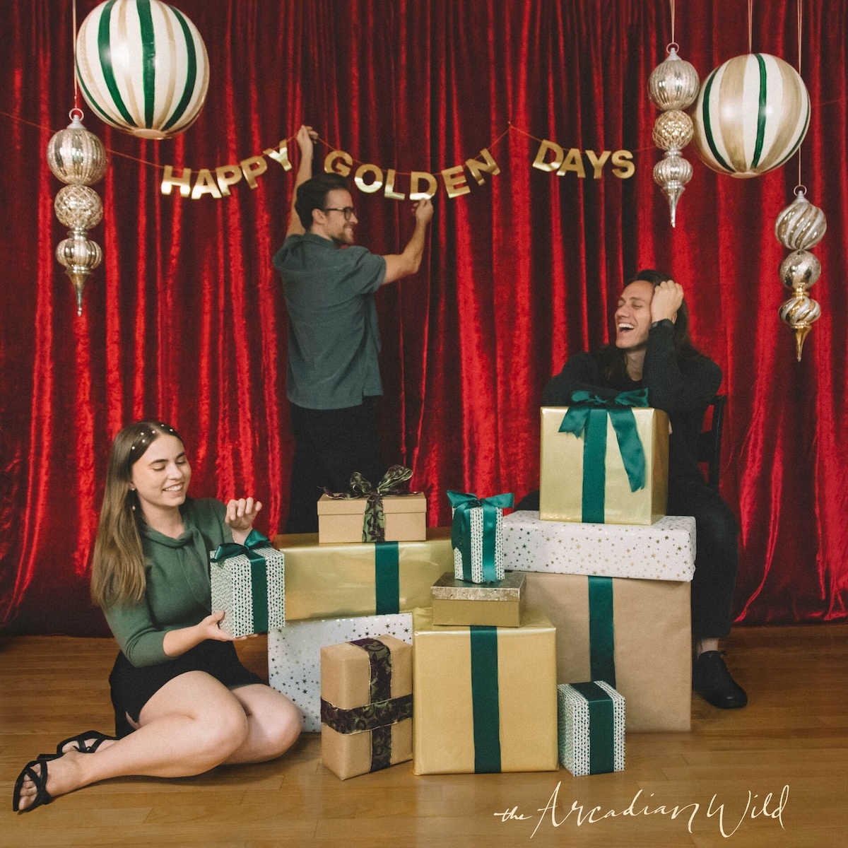 Album Art of The Arcadian Wild's Happy Golden Days EP - A picture of three people in a room with a wooden floor and a bright red curtain in the back. On the curtain, the words 'Happy Golden Days' are hung from a string in gold lettering, and it looks like one man is currently hanging them up. A number of globes and ornaments hang from the ceiling on either side. In the center, in front of that man is a large pile of presents on the floor, with another man sitting crouched and leaning up against the presents. To the left of the presents, a woman is sitting on the floor and affixing a bow to the present. Printed above the floor is the band name in a flowing, hand-written script.