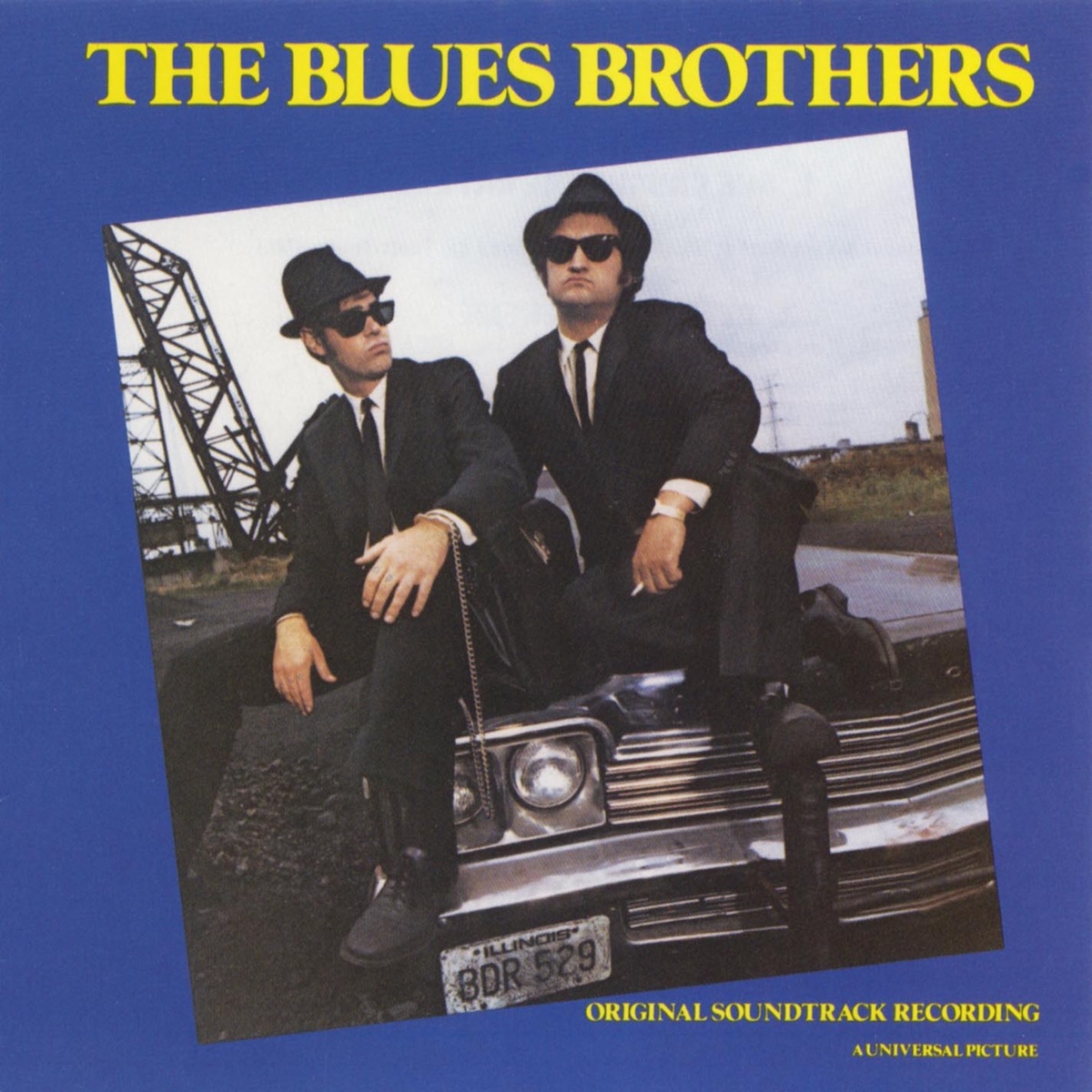 Album Art of The Blues Brothers Soundtrack album - On a solid blue background, a square photo of two men is in color but also rotated about 10 degrees off center counter-clockwise. The photo shows two white men in black suits and pants with thin black ties and tall black hats, also wearing sunglasses. They are sitting on the hood of a black sedan, and a remote place of the Chicago can be seen somewhat in the background, with a gravel/dirt next to the person on the left on the ground and steel railroad bridges in the background. It's a gray, overcast day. At the top in large letters it says the movie name in yellow, plus in much smaller type it says 'Original Soundtrack Recording' and 'A Universal Picture' in the bottom right.