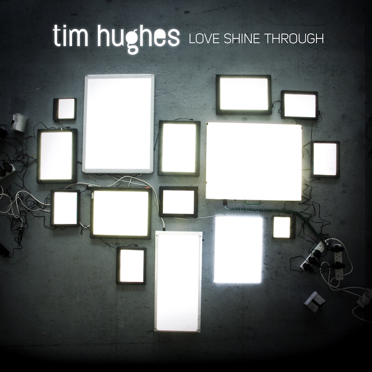 Album Art of Tim Hughes's Love Shine Through album - In a dark room on a carpeted floor, 15 displays of various sizes, from a phone to a tablet to a monitor, are spread across the floor in the approximate shape of a heart. The only light seems to be the displays, which all look to be bright white. Between the devices you can see a bit of carpet, and power cables that are run from power strips to each device. At the top, in large, lowercase white letters, it has the artist's name on the left, with the album cover in much smaller, thin, uppercase letters to the right.