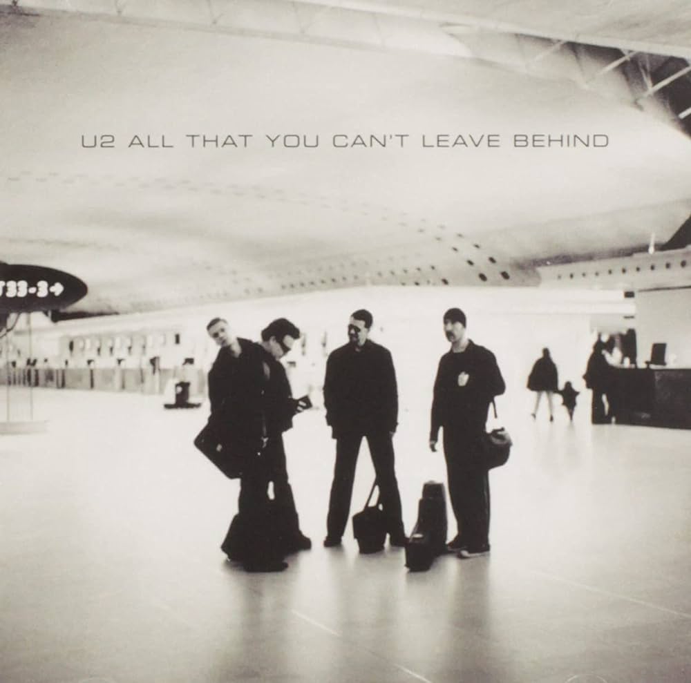 Album Art of U2's All That You Can't Leave Behind album - In a large, open hallway with a tall, curved roof, the four men of U2 stand in the center of the floor with a few bags. It looks like Bono is looking down at a book in his hand, and Larry is looking at Bono. The Edge is standing to the right looking off to left of fhe frame, and Adam is leaning to the left of Bono looking at the camera.