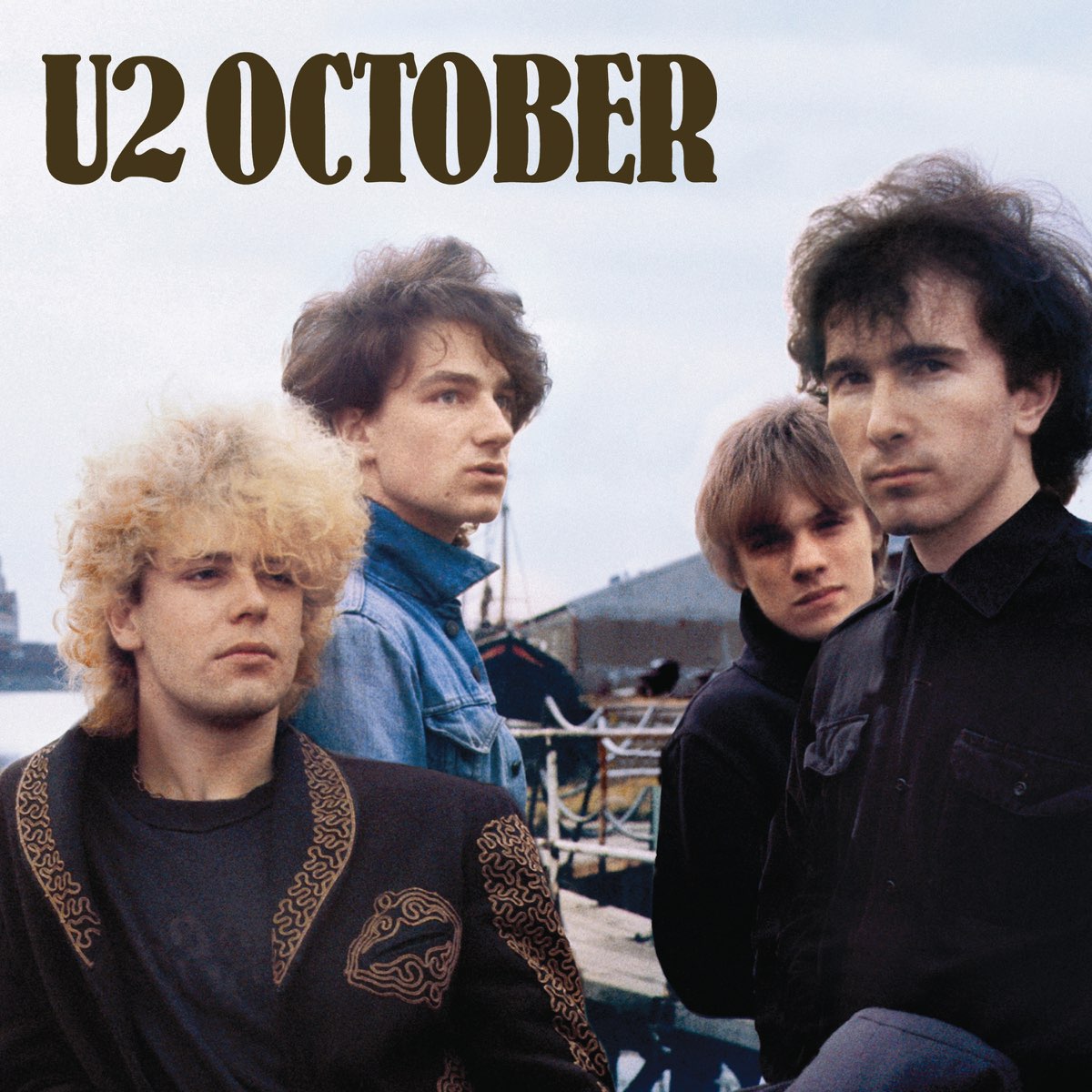  the one on the left is blonde and curly, the second from the right has light brown hair, and the other two have darker brown, windswept hair. The background looks like some docks or industrial area near the water, from what little we can see. In the top left, the band and album name is printed in large letters.