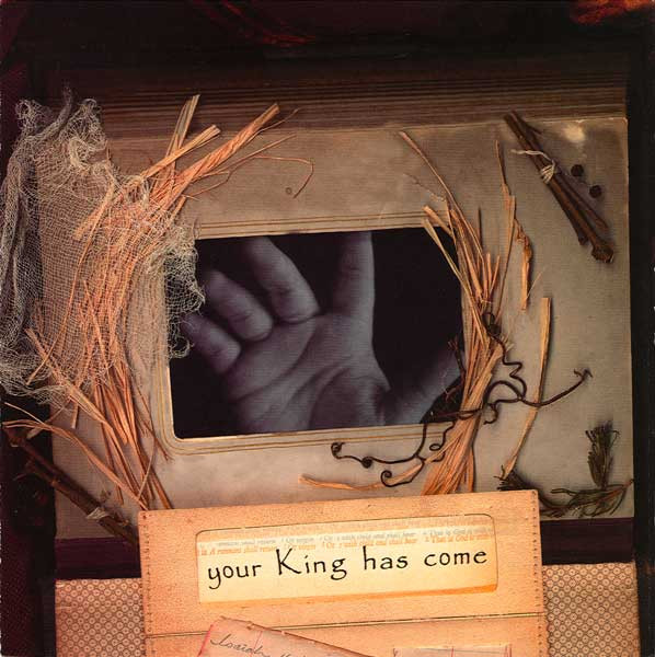 Album Art of the Your King Has Come album - A photo on a dark background of a weathered old book with photos. The photo it is open to is a black-and-white photo of a baby's hand, with a gold double border around the photo. Arranged on the page around the photo are some straw and sticks and some other other items. Printed on a card laid on top of the page is the words 'your King has come', and there is shown some other handwritten pages nearby.