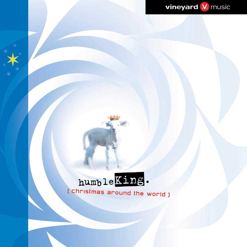 Album Art of Vineyard Music's Humble King album - On a white background, to the left of center, is a white lamb wearing a golden crown and looking at the viewer. Around him is a swirling, circular pattern in grays. On the left side, a blue line is overlaid with a bright, golden star and smaller, blue stars around that. Underneath the lamb is the title 'Humble King' in a ragged typewriter font, then 'Christmas around the world' printed in red and enclosed in brackets. In the top right, a black bar with the 'Vineyard music' logo in white and red.