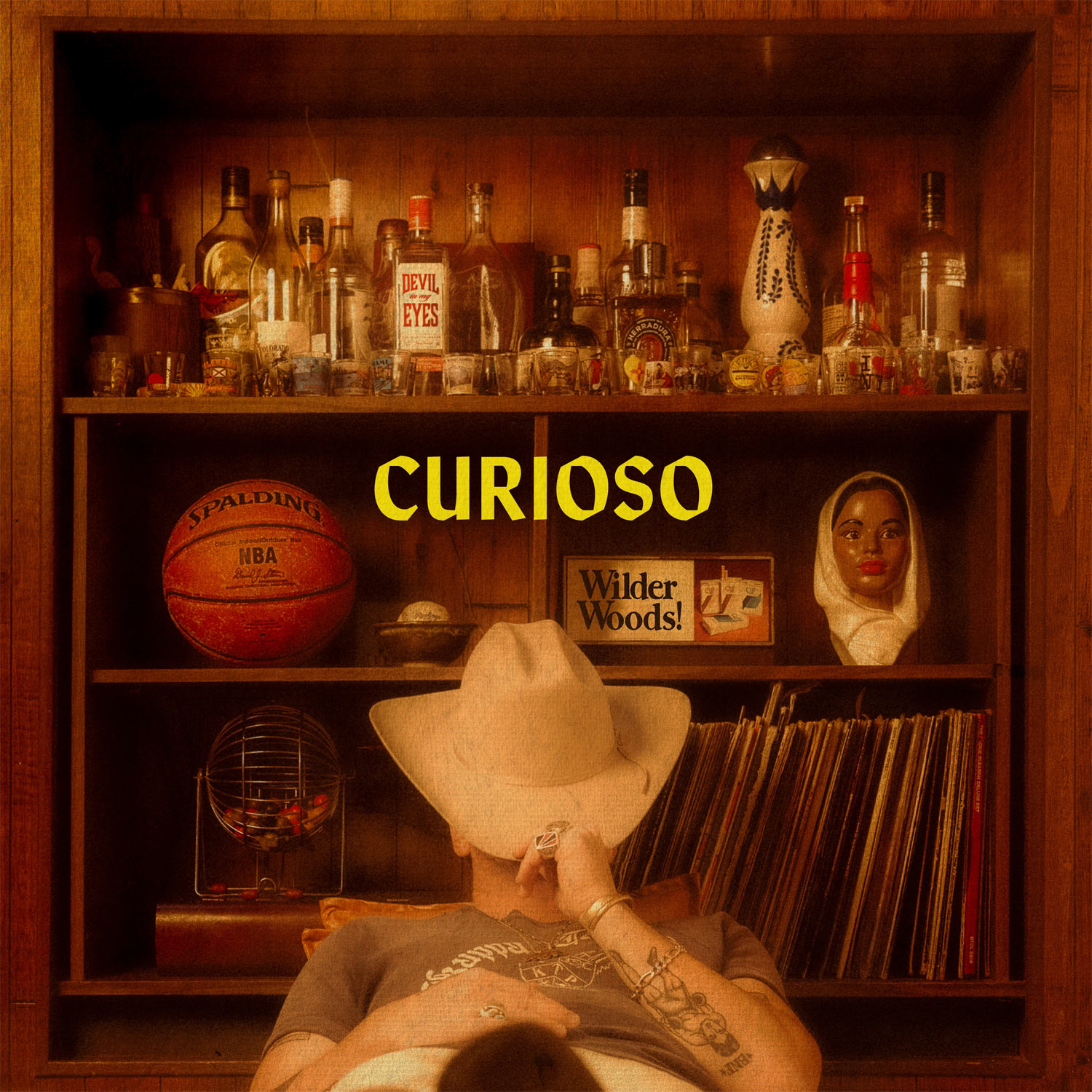 Album Art of Wilder Woods's Curioso album - A photo of a man sitting on a nice chair in front of a bookshelf, though there's no books here. The top shelf is dozens of liquor bottles and dozens of shot glasses. The middle shelf has an NBA basketball, a baseball in a bowl, and cigarette ad that says 'Wilder Woods!', plus a bust that looks like a dark-skinned woman with a head covering. On the last shelf, there's about 30 records, maybe.</body></html>