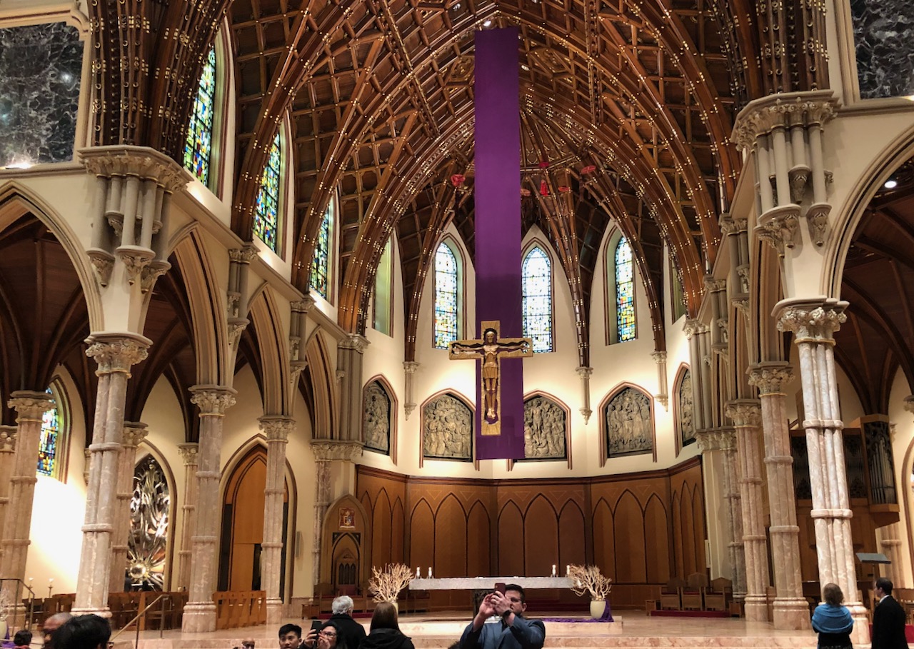 Photo of the interior of a Roman Catholic Church, with an ornate cross above an altar in the center surrounded by beautiful columns and topped with an arched roof