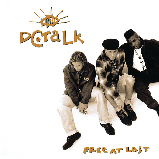 Album Art of dc Talk's Free At Last album - On a white background, a black-and-white photo of three young men from above. A long-haired white man on the left, a short-haired man with a black hat on in the middle, and a black man with lots of dark hair sticking up on top but none on the sides and wearing multple crosses around his neck. The band name is in the top left, with an eye illustration above it with a cross inside the eye. At the bottom right below the photo is the album name.